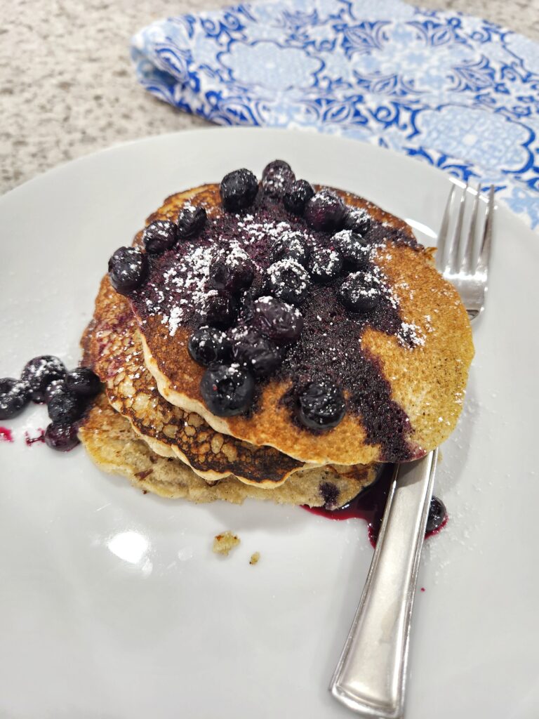 Barley pancakes with a fork stacked on white plate, topped with blueberries and sugar beside and a blue and white napkin.