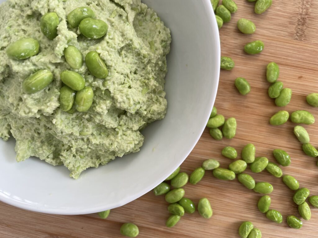 High protein hummus presented in white bowl on wood cutting board surrounded by edamame beans.