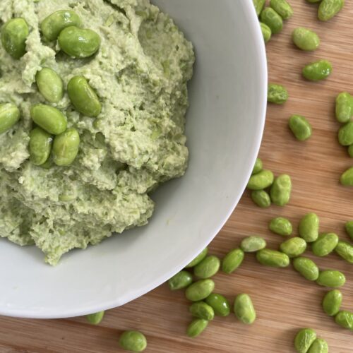 High protein hummus presented in white bowl on wood cutting board surrounded by edamame beans.