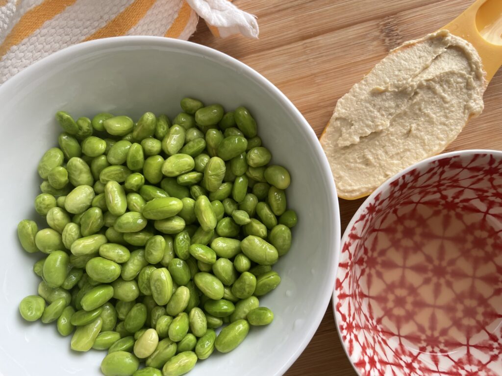 Edamame beans in white bowl, lemon juice in red bowl and hummus in yellow measuring cut on wood cutting board.