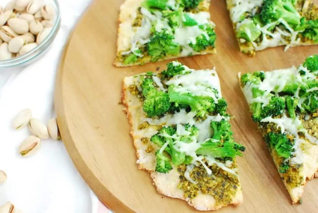 An image of pistachio pesto pizza cut in slices, on top of a wooden board.