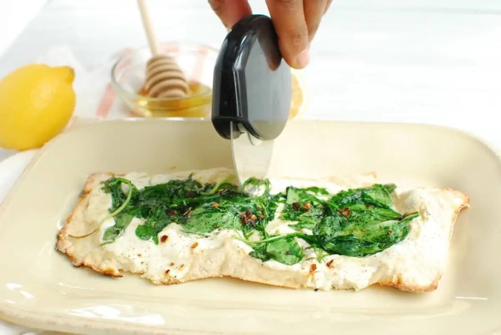 Lemon ricotta pizza topped with spinach on a dish, being cut with a pizza cutter.