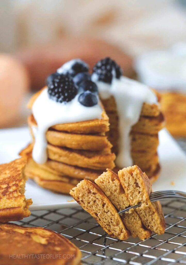 A stack of sweet potato pancakes topped with berries and yogurt. The front of the image shows part of the stack of pancakes on a fork. 