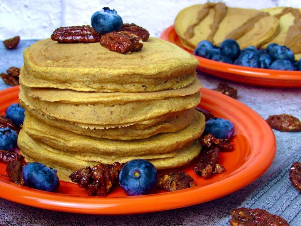 A stack of sweet potato pancakes on an orange plate, topped with candied pecans and blueberries. 