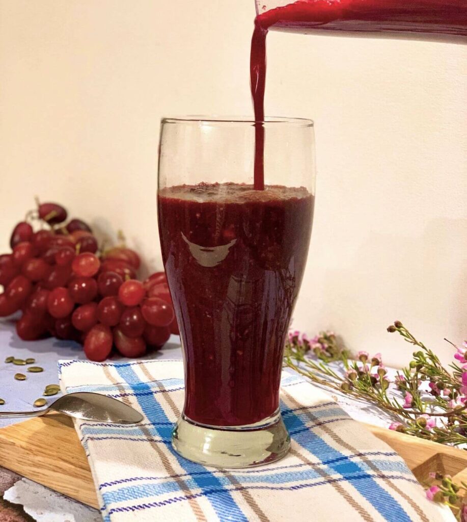 An image of a beet smoothie being poured into a glass. The glass is on top of a checkered tea towel, with grapes and flowers in the background. 