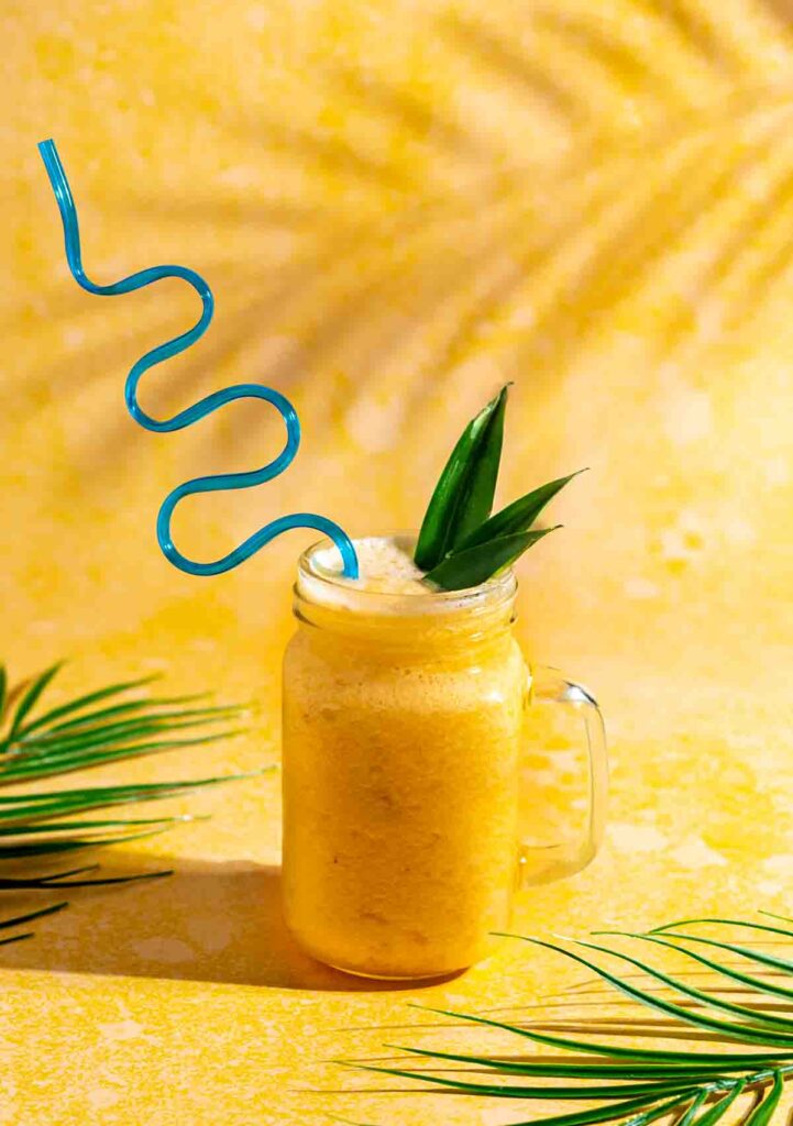 Pineapple slushie in a glass jar with a  blue crazy straw. The background is yellow.
