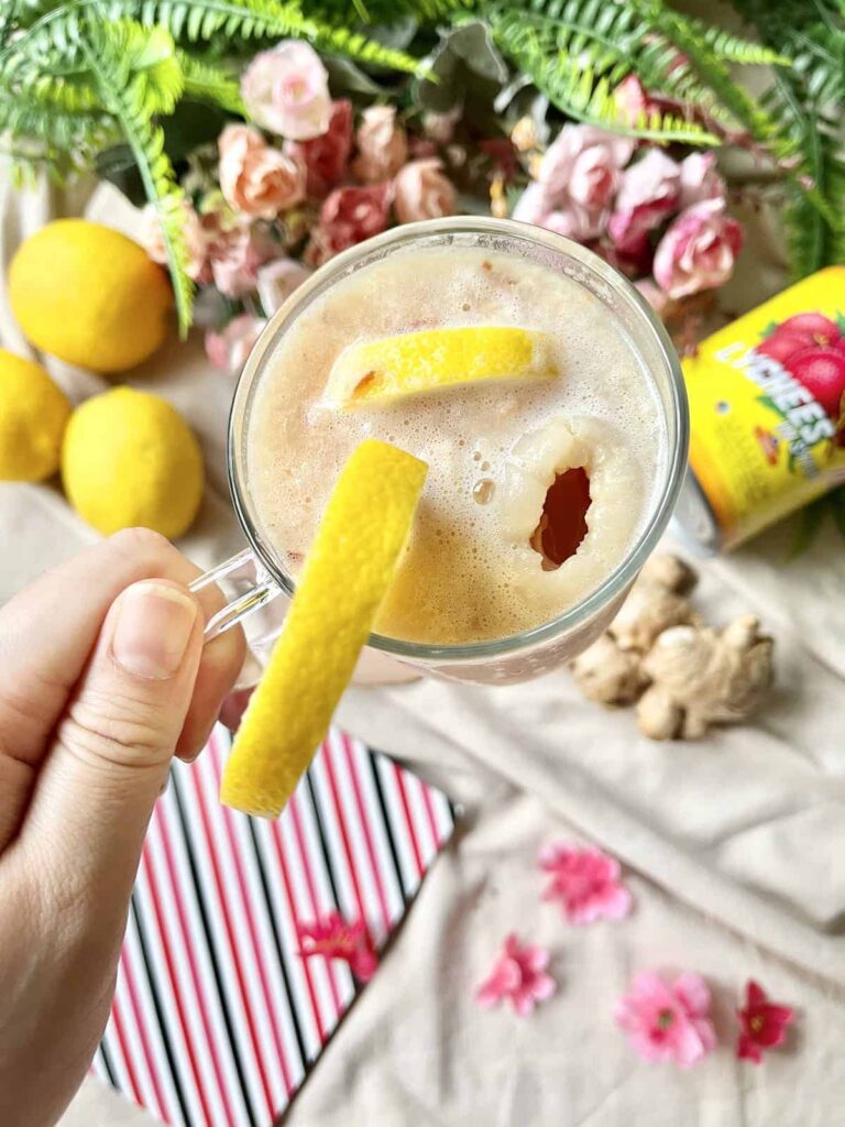 Lychee juice photographed in a glass being held. In the background is flowers, lemons, and lychees. 