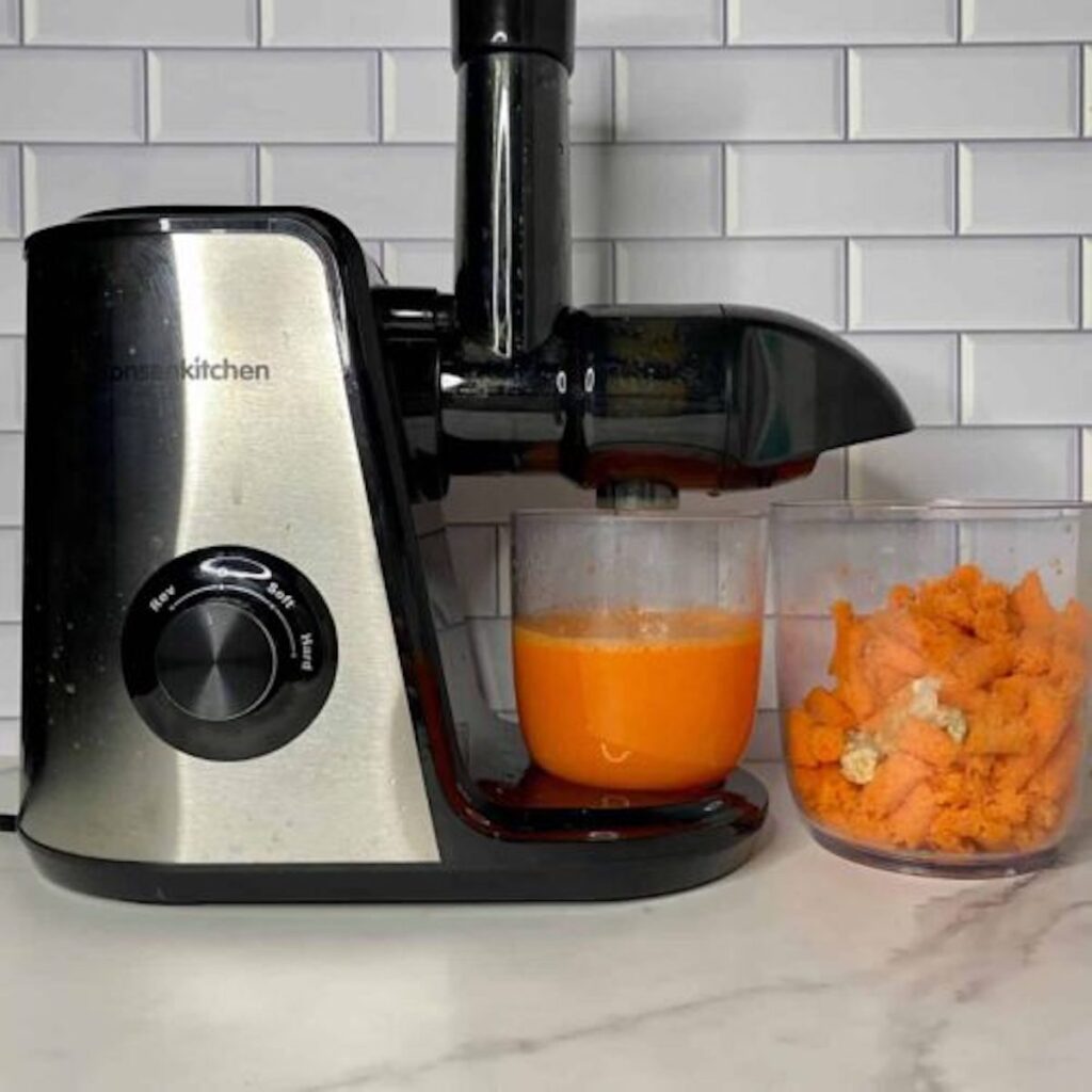 Carrot orange ginger juice in a dish with a juicer. Next to the juice is a dish with leftover carrot and ginger. 