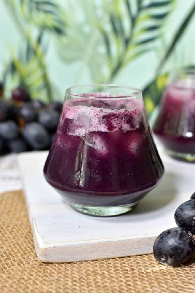 Grape juice in a glass with ice. Grapes are surrounding the glass of juice in the background. 