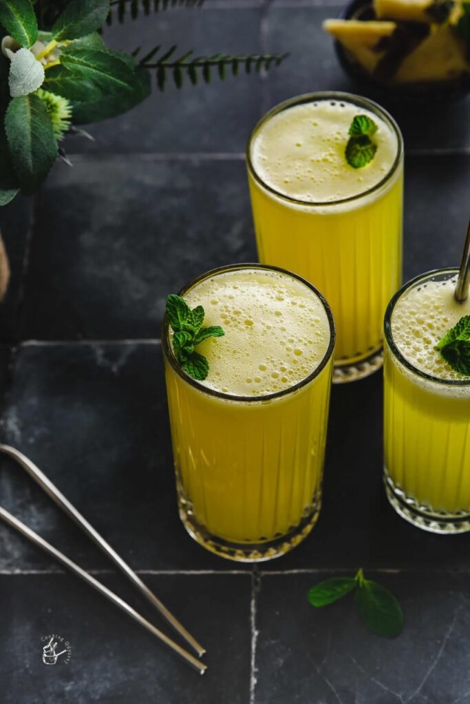 3 tall glasses of pineapple juice, topped with fresh mint. In the background is a dish with sliced pineapple. 