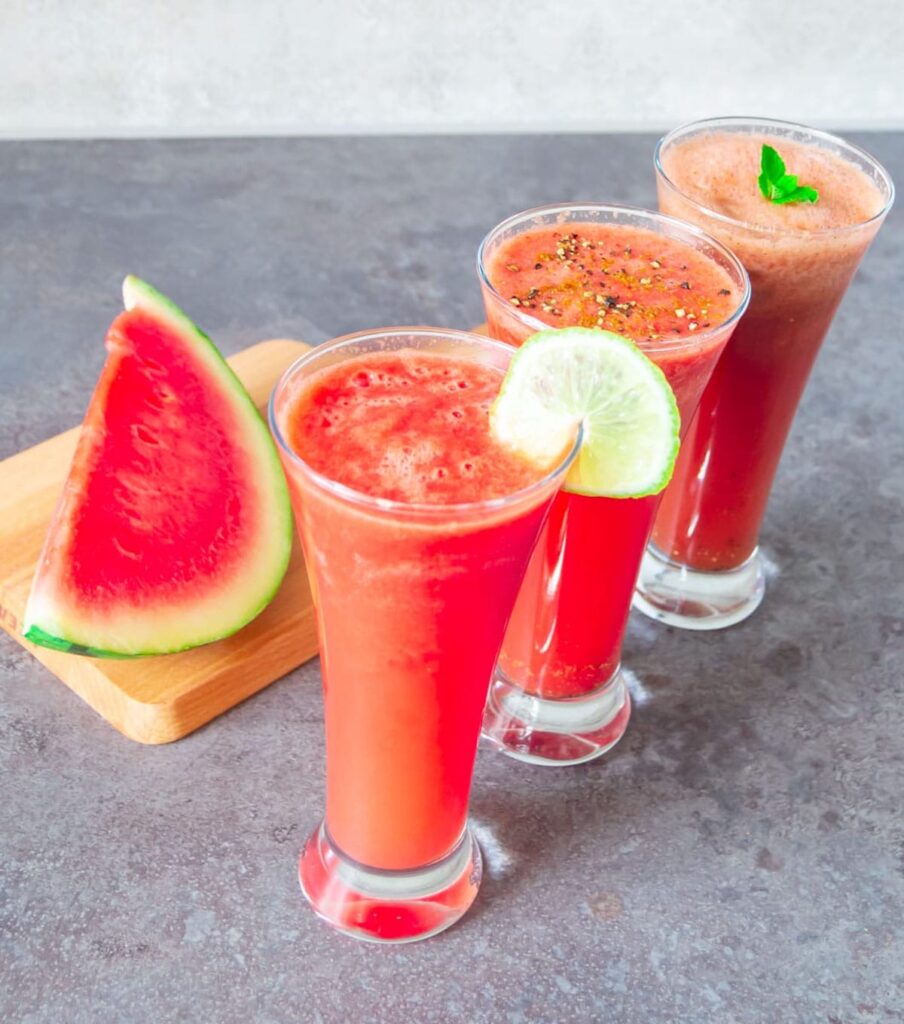 3 glasses of watermelon juice lined up, with lime on the rim of one glass.  A slice of watermelon sits on a wooden cutting board next to the watermelon juice. 