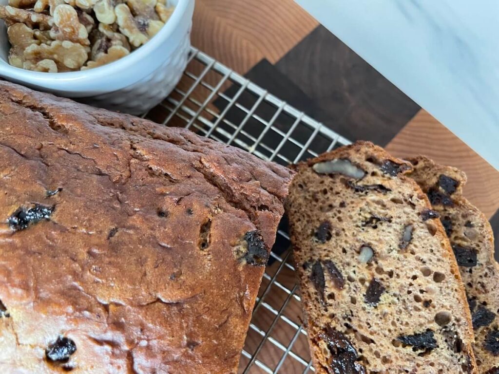 An image of prune banana bread on a cooling rack, with two slices cut off. A white dish with walnuts is next to the loaf.