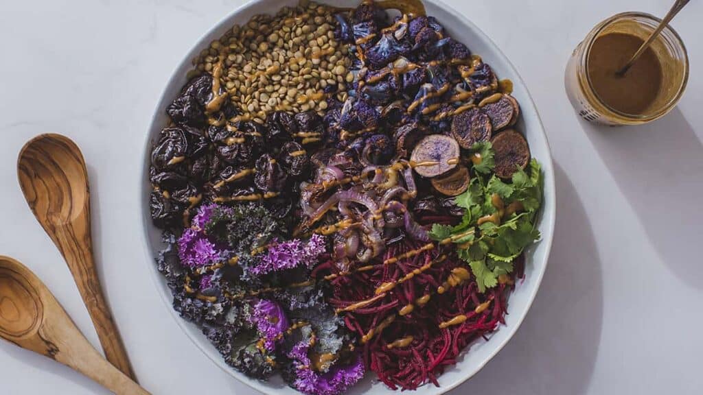 A spiced purple power bowl filled with prunes, fresh herbs, purple kale, broccoli, beets and lentils in a white bowl. Next to the bowl is a jar of dressing and wooden salad spoons.