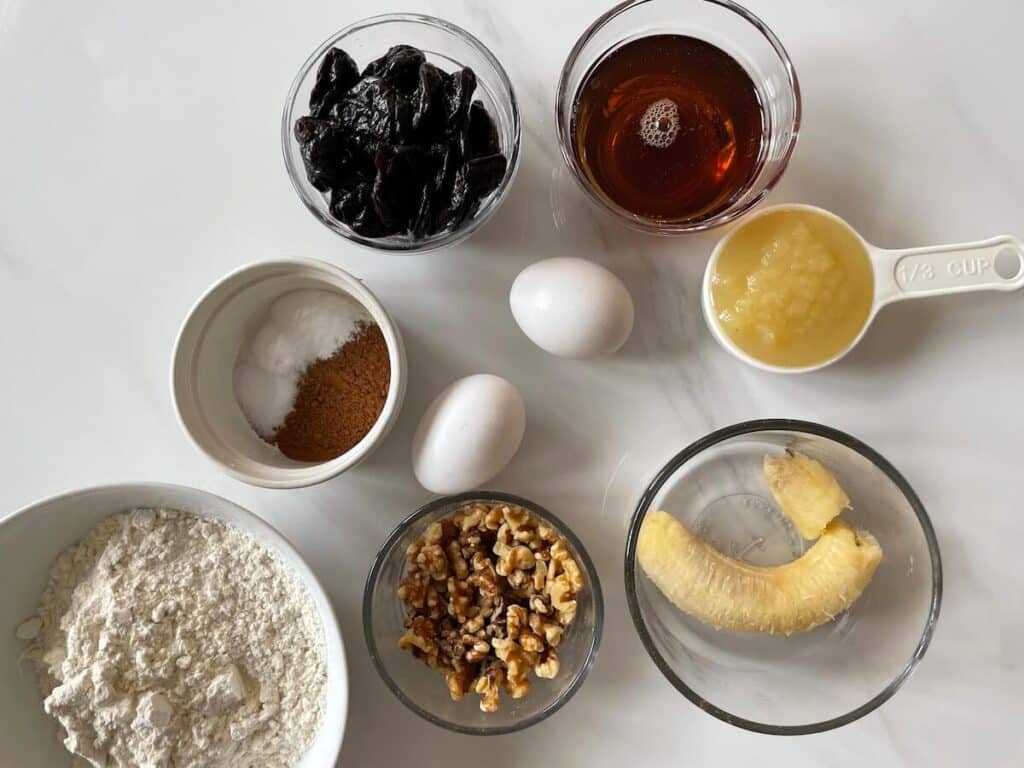 Prune banana bread ingredients, including banana, walnuts, eggs, cinnamon, applesauce, prunes, honey, and flour.