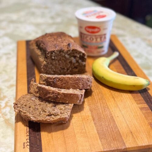 Banana ricotta bread sliced on a wooden cutting board, next to a container of ricotta and a banana.