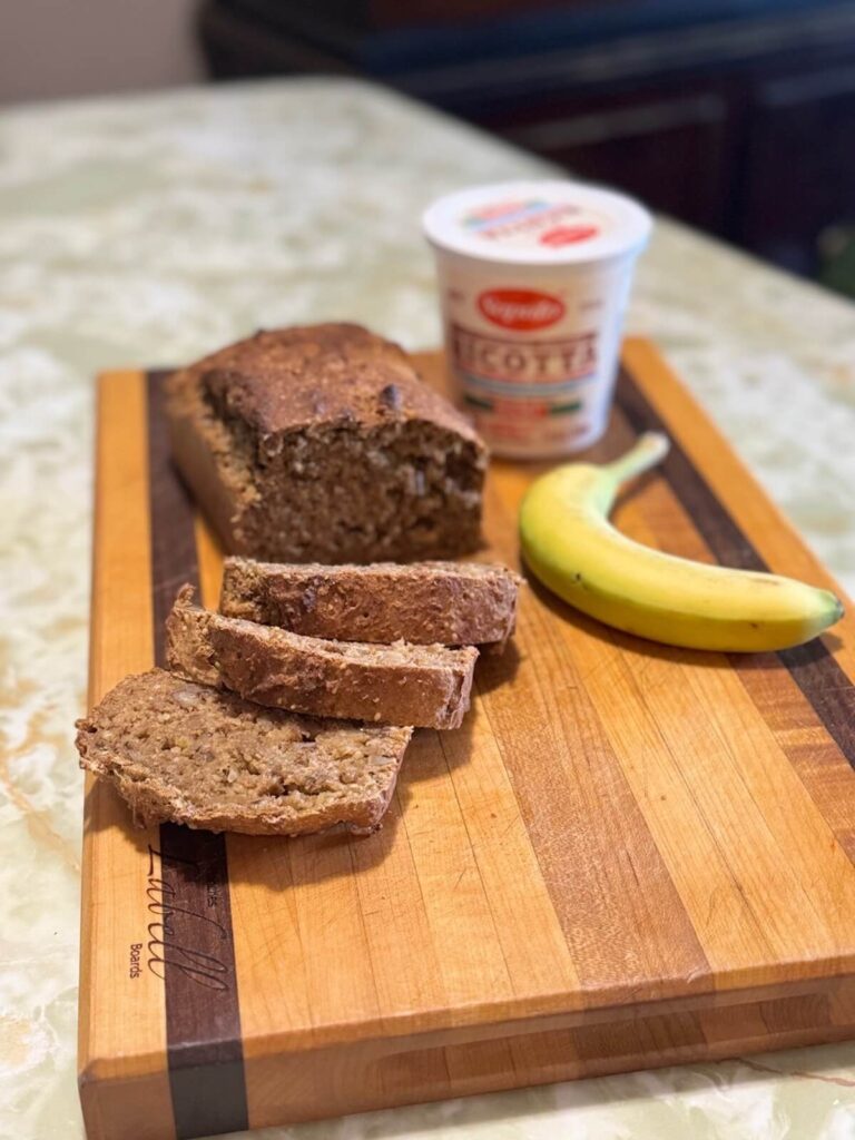 Banana ricotta bread sliced on a wooden cutting board, next to a container of ricotta and a banana.