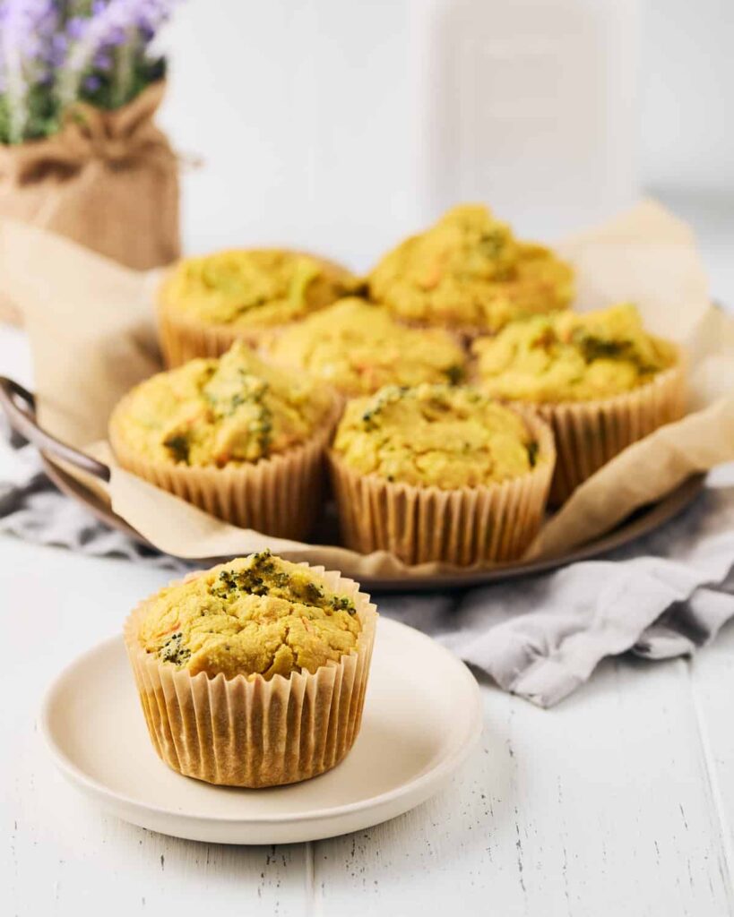 Broccoli cheddar egg cups made with chickpea flour in muffin form. One is placed on a plate, and a few on parchment paper in the background.