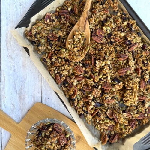 Fruit and nut granola presented on parchment paper on a sheet pan with a wooden spoon on top, and beside a wood cutting board with granola on it.