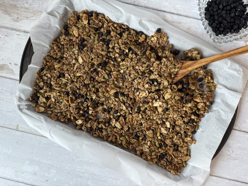 Blueberry granola presented on a sheet pan covered in parchment paper beside a clear bowl of dried blueberries.