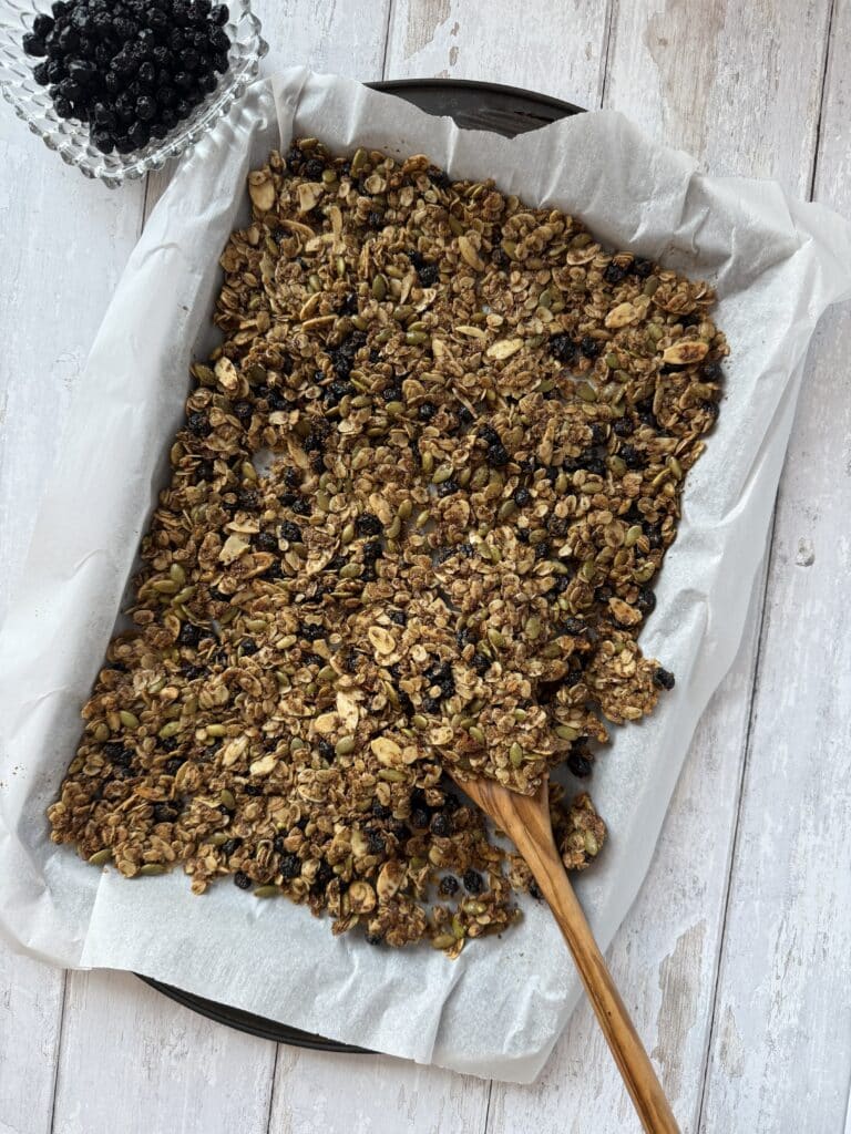 Simple blueberry granola on a sheet pan with parchment paper beside a bowl full of dried wild blueberries.