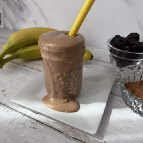 This is banana prune smoothie on a white plate with bananas, prunes and cocoa in the background.