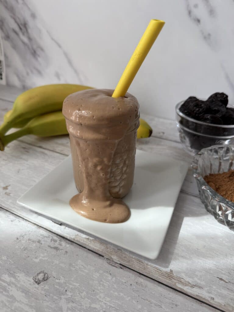 This is banana prune smoothie on a white plate with bananas, prunes and cocoa in the background.