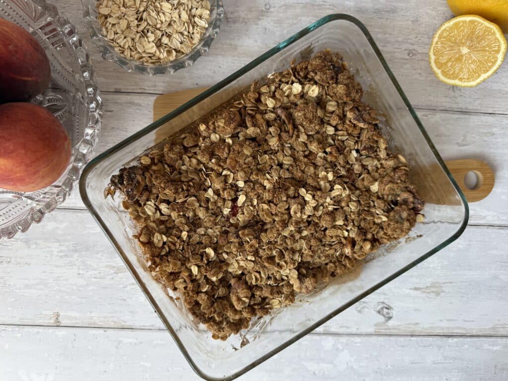 Peach oat crisp in a glass container surrounded by oats, peaches and lemons.