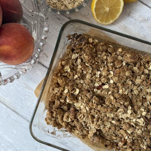 Peach oat crisp in a glass container surrounded by oats, peaches and lemons.