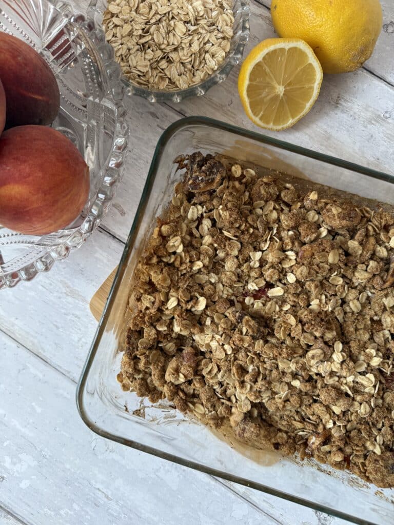 Peach oat crisp in a glass container surrounded by oats, peaches and lemons.