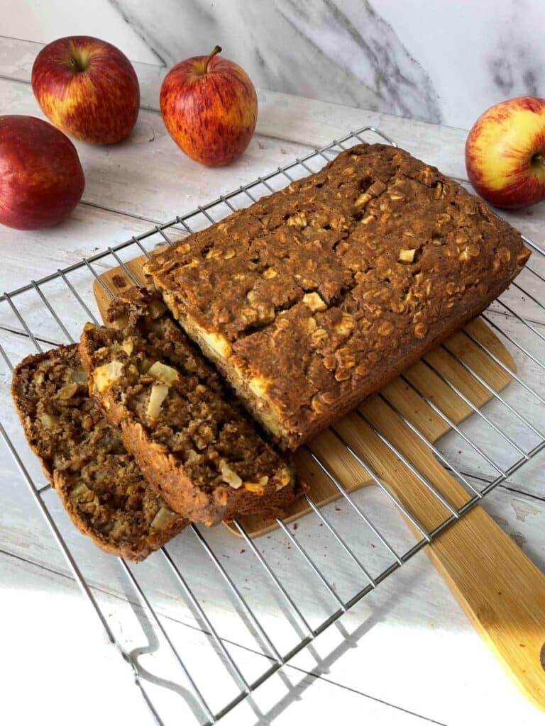 Apple and cinnamon loaf cake presenting on cooling rack on top of wood cutting board surrounded by whole apples.