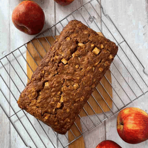 Apple and cinnamon loaf cake presenting on cooling rack on top of wood cutting board surrounded by whole apples.