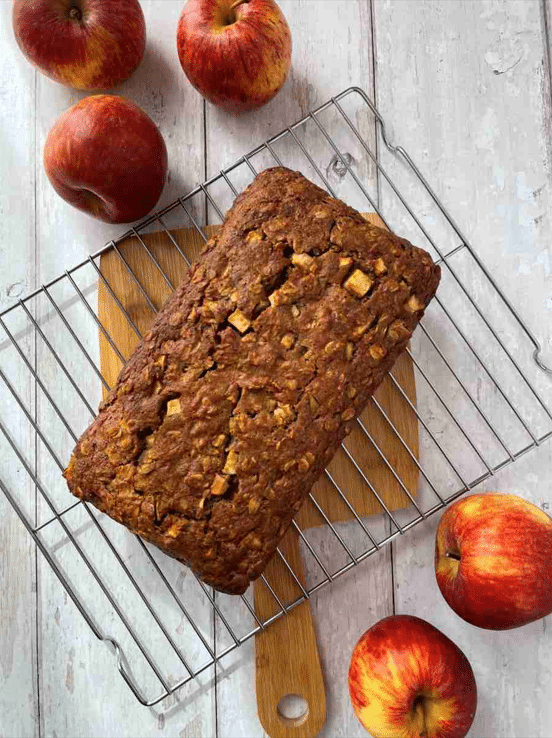 Apple and cinnamon loaf cake presenting on cooling rack on top of wood cutting board surrounded by whole apples.