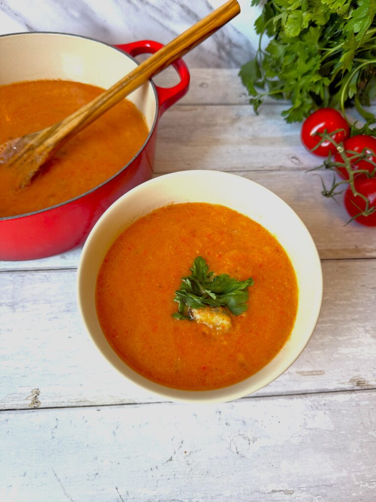 Blended soup in a white bowl on a white picnic table beside a red French filled with more soup and a wooden spoon. The soup is sitting beside tomatoes on a vine and a bunch of parsley.