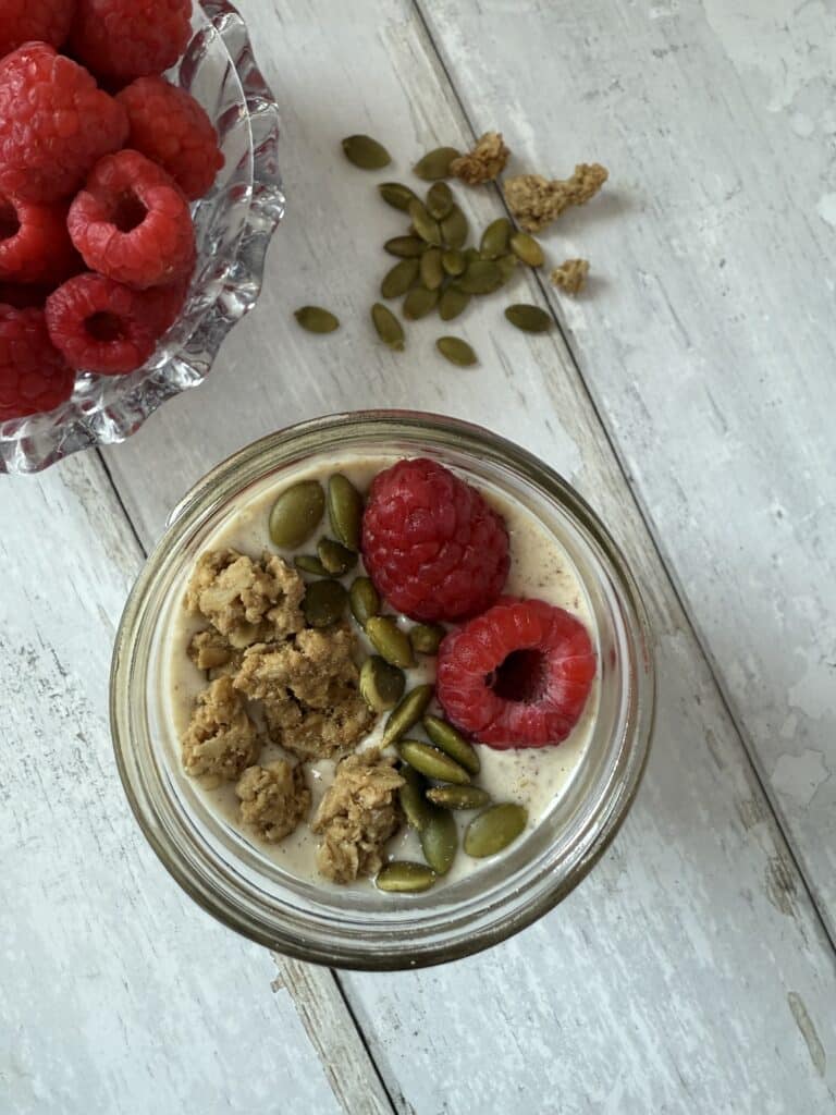 Blended overnight oats topped with granola, pumpkin seeds and fresh raspberries beside a bowl of raspberries.