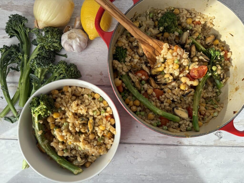 Mussel risotto presented in red french oven with wooden spoon, beside white bowl filled with risotto beside a lemon, onion, garlic and broccolini.