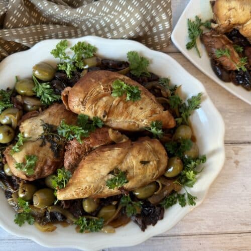 Moroccan chicken with prunes on a white serving platter beside a smaller plate of chicken and displayed on top of a brown tea towel.