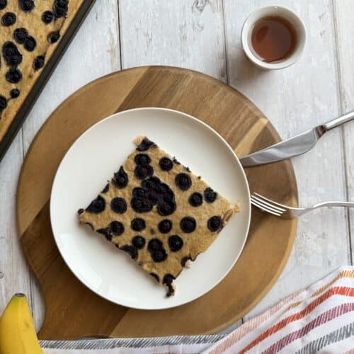 Sheet pan tofu pancake with a slice of high protein pancake on white plate, sitting on wood cutting board beside a banana and maple syrup.