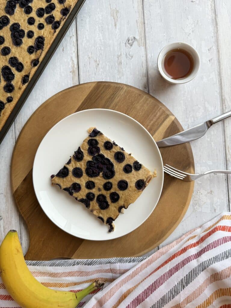 Sheet pan tofu pancake with a slice of high protein pancake on white plate, sitting on wood cutting board beside a banana and maple syrup.