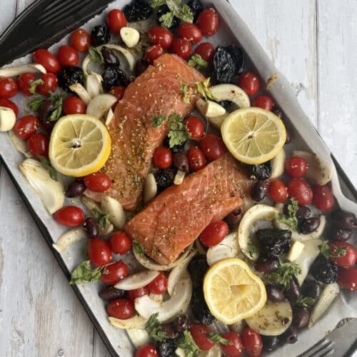 Salmon fillets, lemon, prunes, tomatoes, and olives on a sheet pan covered in parchment paper.