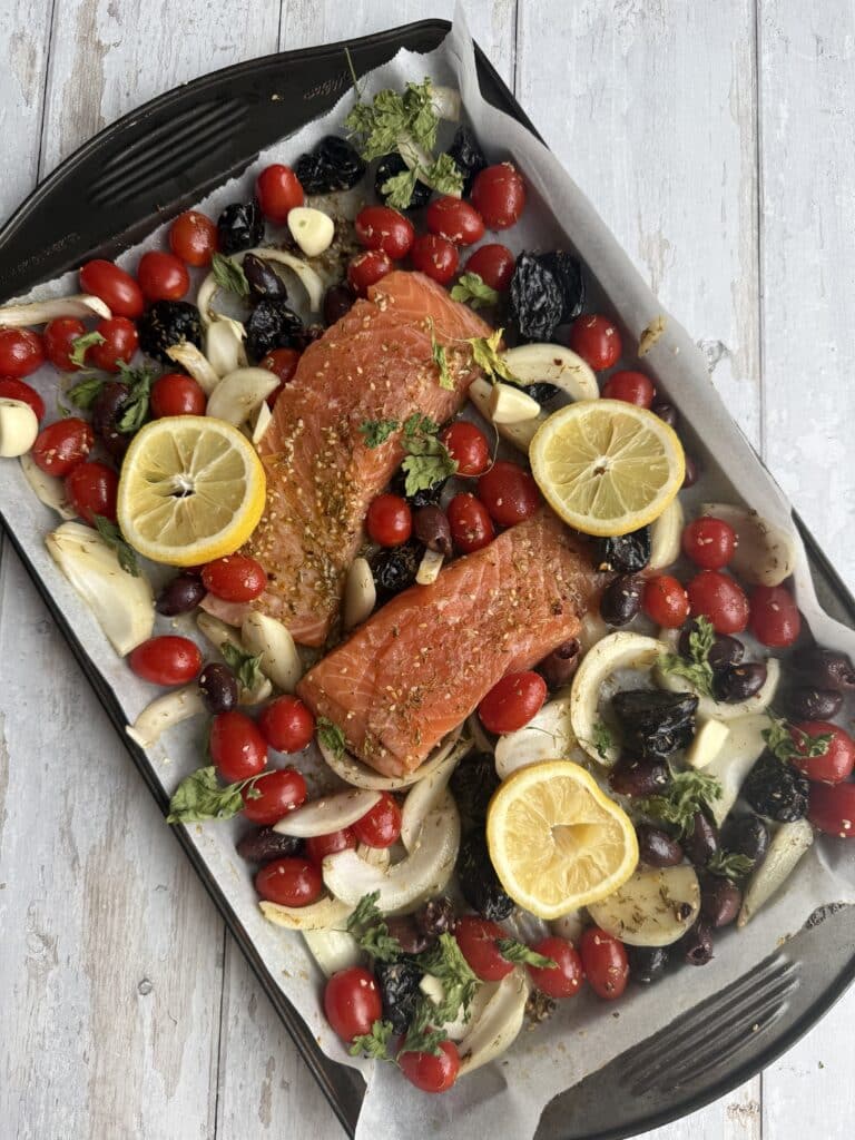 Salmon fillets, lemon, prunes, tomatoes, and olives on a sheet pan covered in parchment paper.