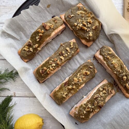 Six walnut crusted salmon fillet presented on parchment paper beside lemon, garlic, walnuts and dill.