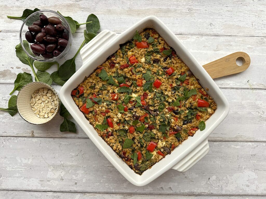 Mediterranean oatmeal in a white dish beside a wooden spoon, oats and olives.