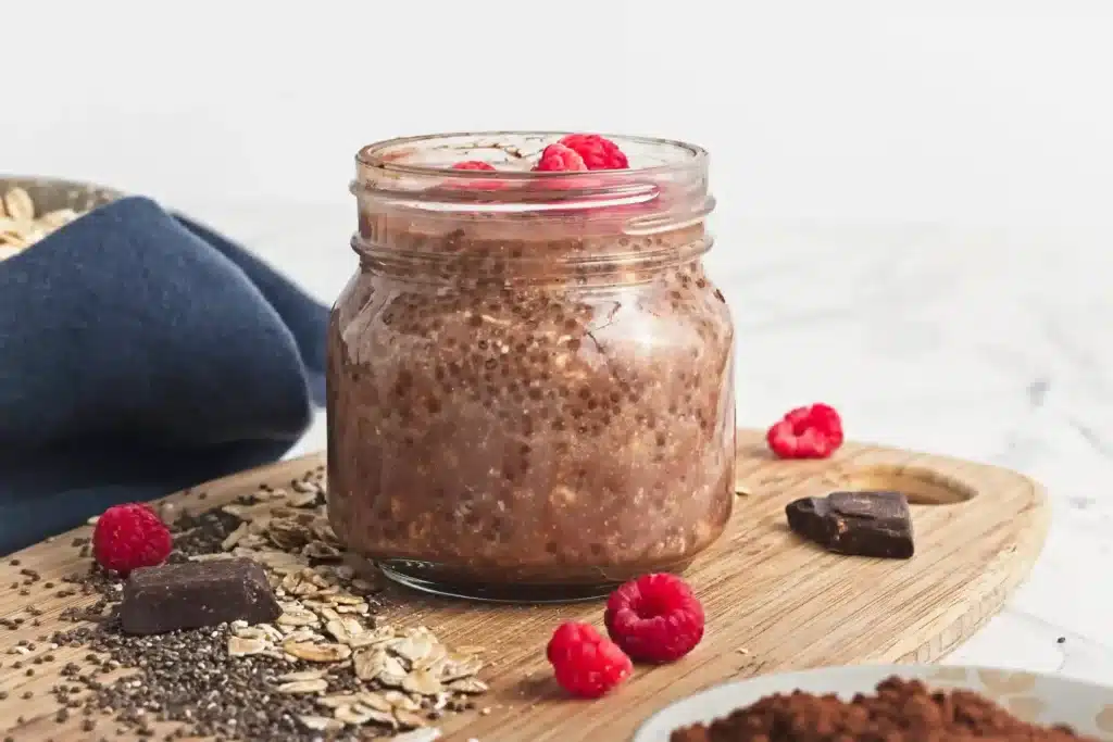 Chocolate overnight oats in a glass jar, topped with fresh raspberries, with chocolate chunks and oats scattered on a wood board.