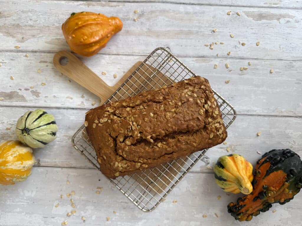 Top down view of a healthy pumpkin bread loaf surrounded by gourds and displayed on a wood cutting board.