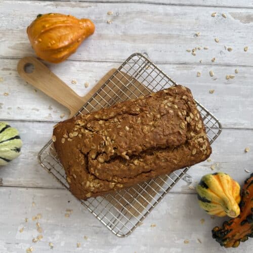 Top down view of a healthy pumpkin bread loaf surrounded by gourds and displayed on a wood cutting board.